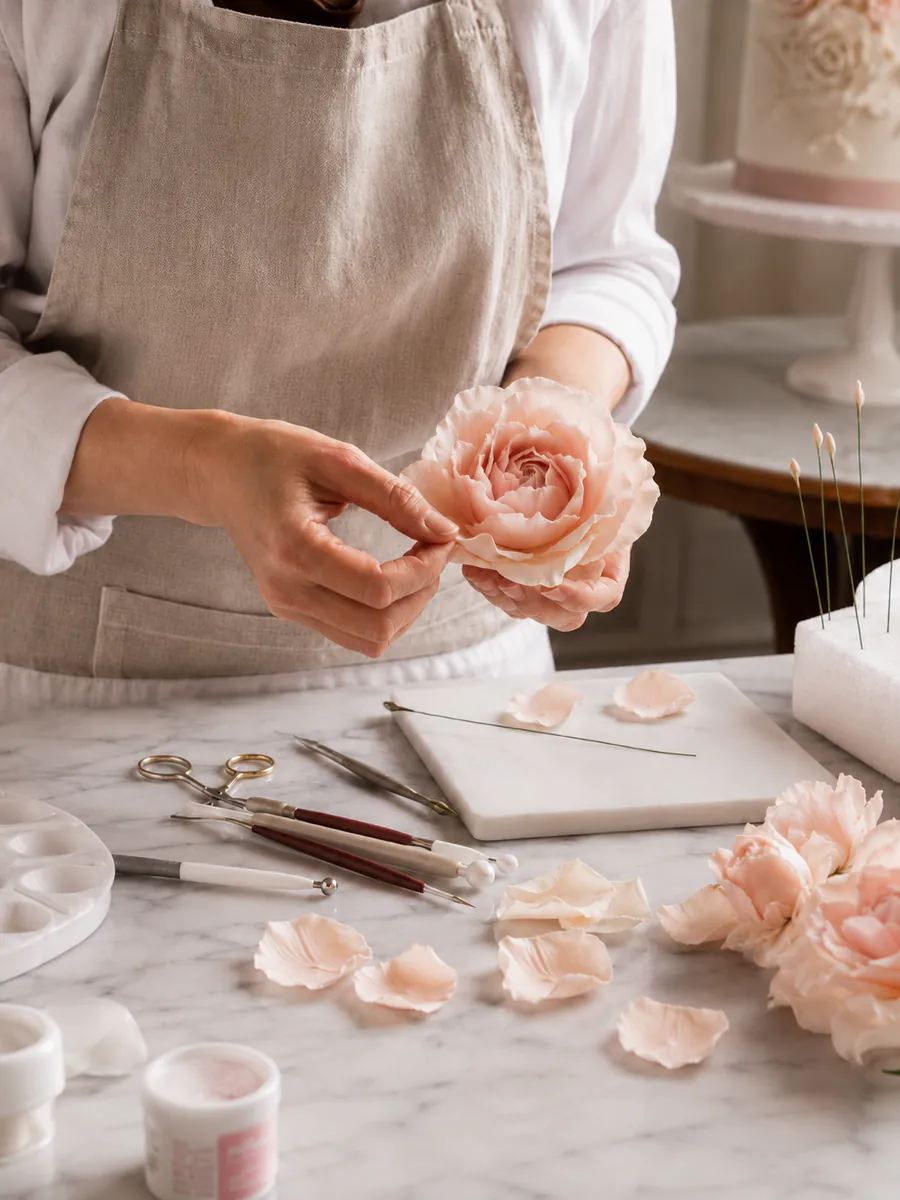 Hände formen eine Zuckerrose aus Blütenpaste, Modellierwerkzeug und Blütenblätter auf der Arbeitsfläche